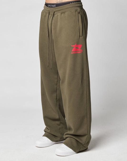 Person wearing LIVE FAST DIE YOUNG Live Fast Sweatsuit Jogger in loose, olive green with Oversize Fit and pink graphic logo on the left thigh, paired with white sneakers and a black LIVE FAST waistband. Plain light background.