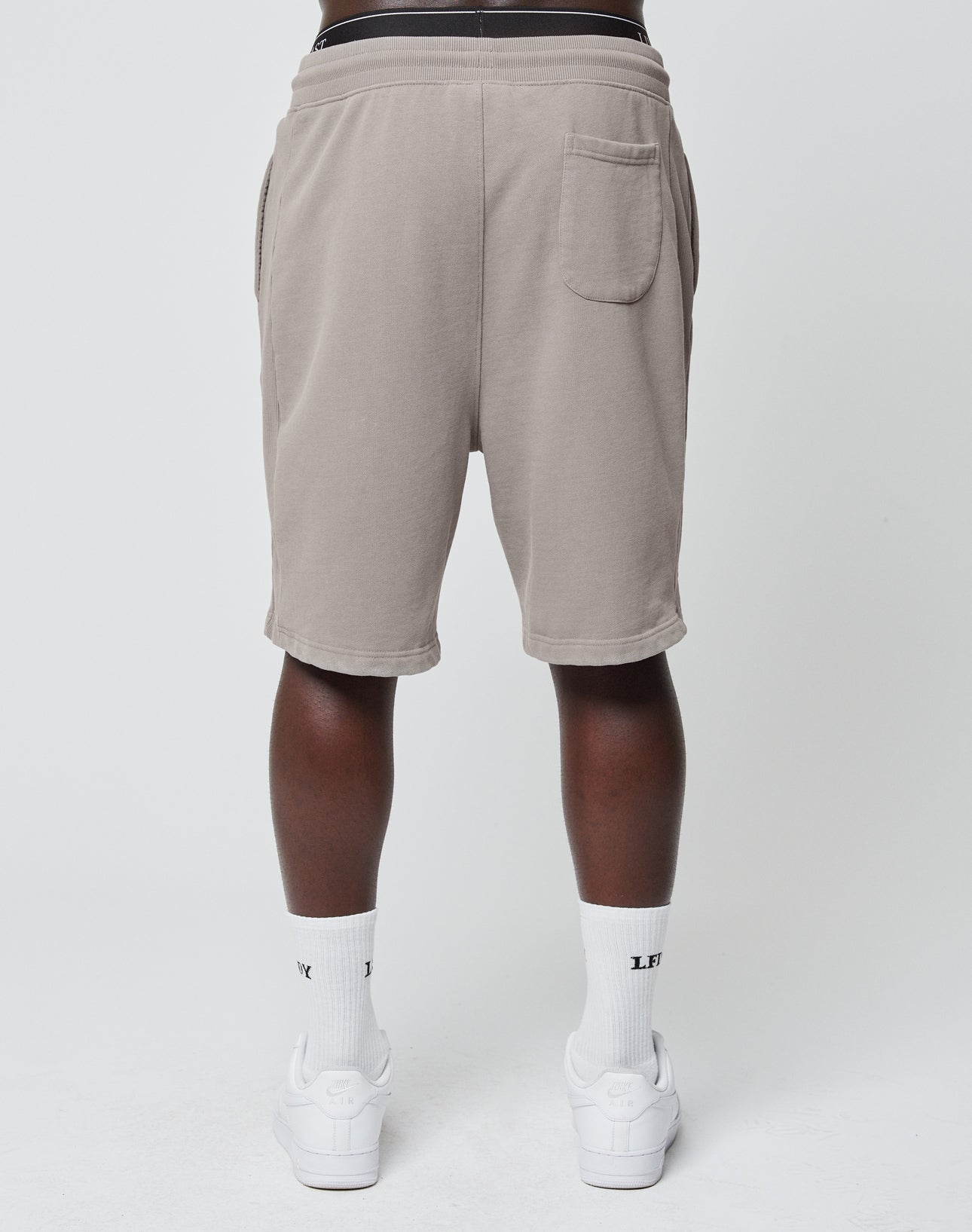 A person stands with their back to the camera, wearing LIVE FAST DIE YOUNG Basic Heavy Shorts in light gray Regular Fit, featuring a single right back pocket, paired with white socks and sneakers.