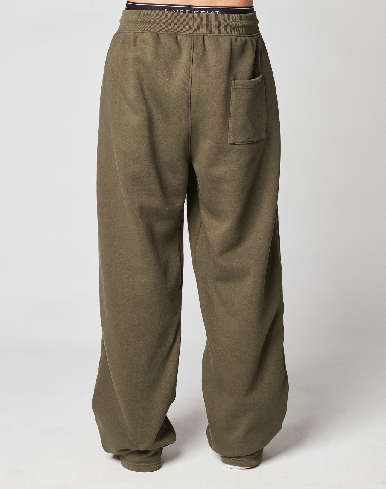 A person wears the LIVE FAST DIE YOUNG Live Fast Sweatsuit Jogger—loose-fitting olive green pants with an oversize fit and visible back pocket—posing against a light background, shown from the waist down.