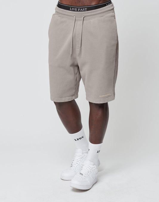 A person wearing LIVE FAST DIE YOUNG Basic Heavy Shorts in beige, featuring a black waistband with LIVE FAST text, plus white LFDY logo-print socks and white sneakers stands against a plain white background.