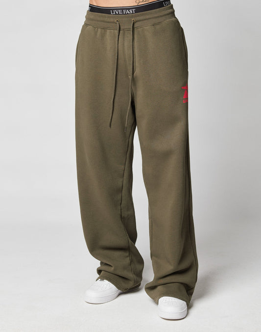 Wearing the LIVE FAST DIE YOUNG Live Fast Sweatsuit Jogger in olive green, featuring an oversize fit, wide elastic waistband with LIVE FAST text, drawstring, a red graphic on the left thigh, and paired with white sneakers.