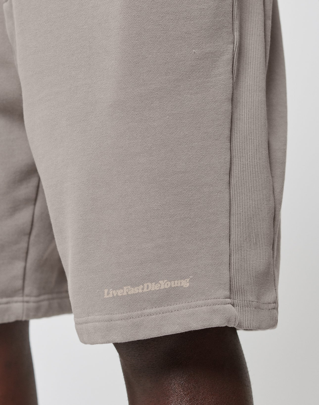 A close-up of a person wearing LIVE FAST DIE YOUNGs Basic Heavy Shorts in light brown, featuring a subtle LiveFastDieYoung logo near the hem and crafted for a comfortable regular fit.