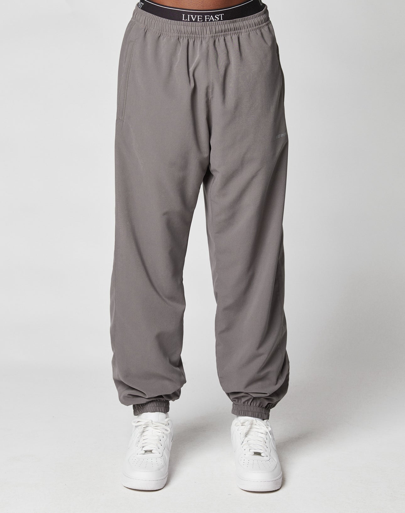 A person models LFDY Trackpants by LIVE FAST DIE YOUNG—gray joggers with an elastic waistband reading LIVE FAST, elastic cuffs, and a regular fit—paired with white sneakers against a plain white background.
