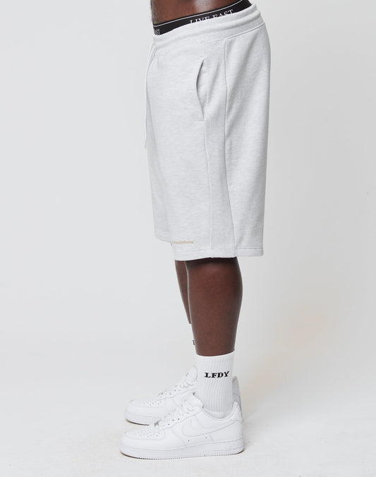A person stands sideways wearing light gray LIVE FAST DIE YOUNG Basic Heavy Shorts with an elastic waistband, white crew socks featuring the LFDY logo, white Nike Air Force 1 sneakers, and a visible black waistband against a plain white background.