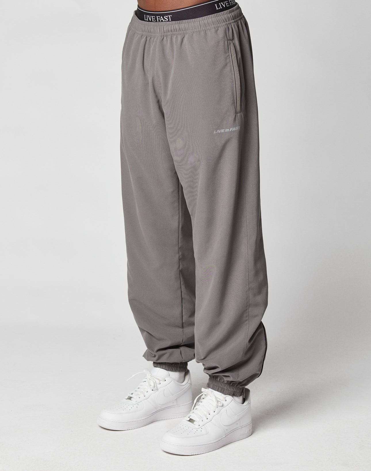 A person wears LFDY Trackpants by LIVE FAST DIE YOUNG—loose-fitting gray joggers with Regular Fit and LIVE FAST reflective print near the pocket and elastic waistband—paired with white sneakers against a plain, light background.