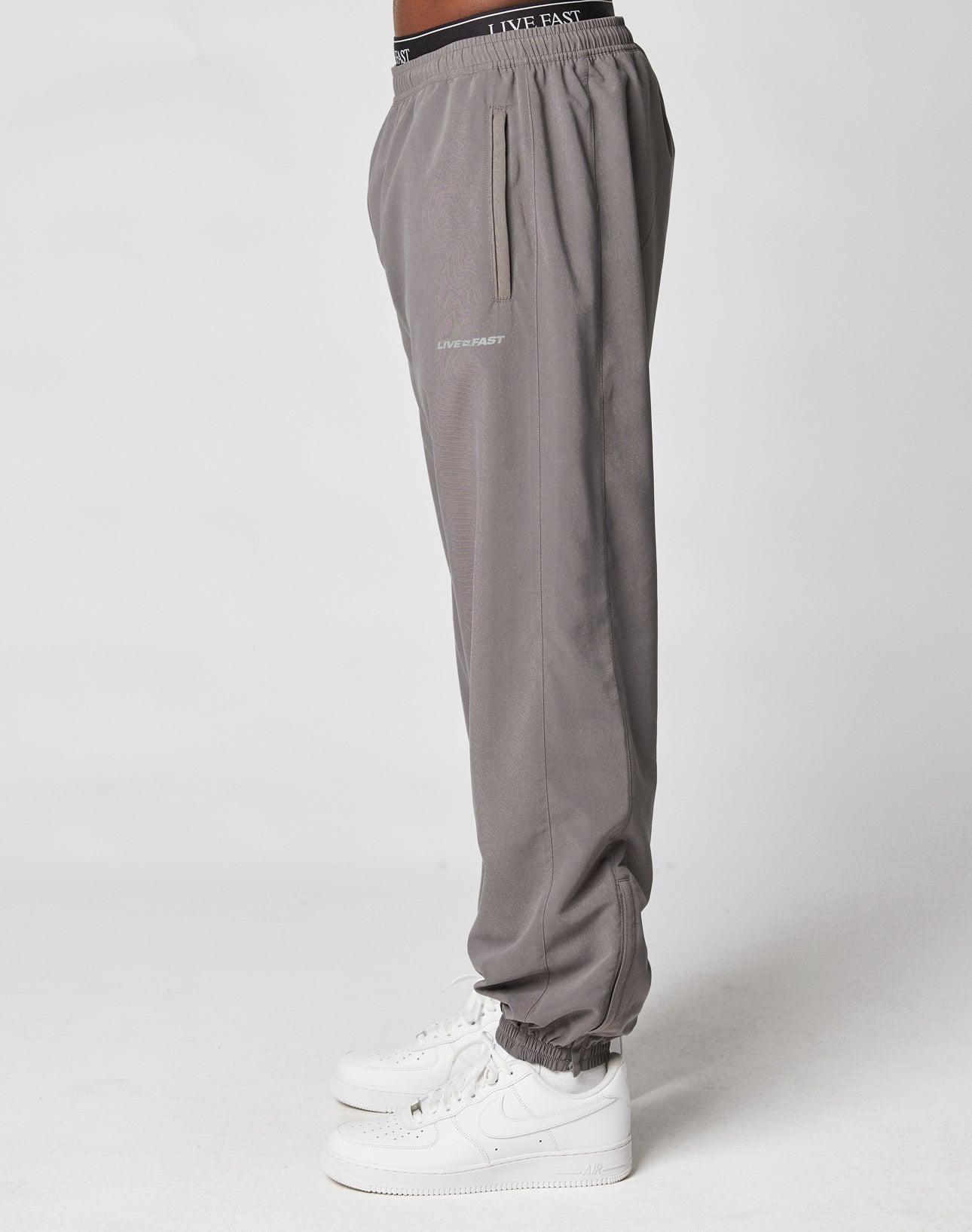 A person stands sideways in LFDY Trackpants by LIVE FAST DIE YOUNG, featuring a Regular Fit and LIVE FAST reflective text on waistband and thigh, paired with white sneakers against a plain white background.