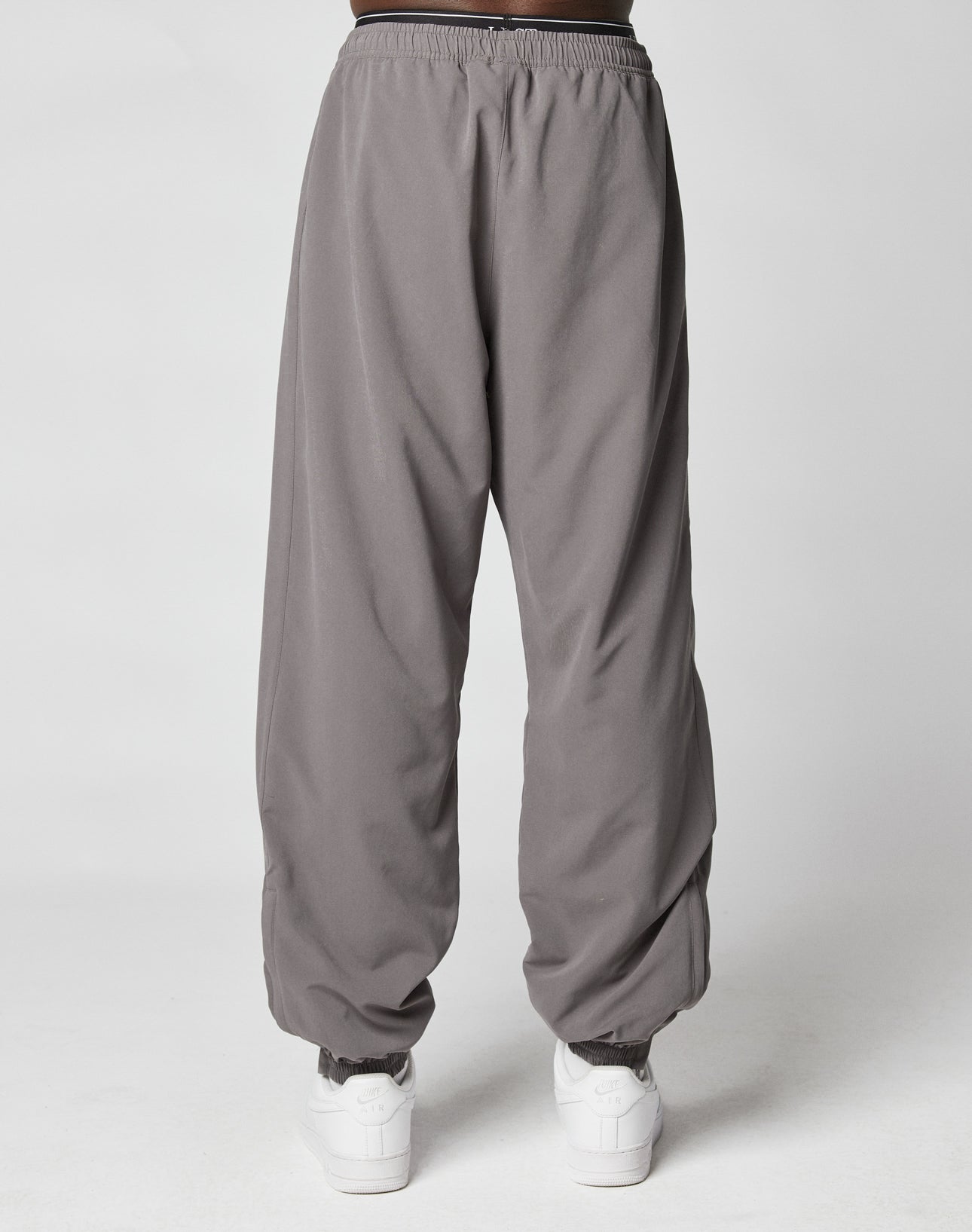 A person wears gray LFDY Trackpants by LIVE FAST DIE YOUNG, featuring an elastic waistband and cuffs, paired with white sneakers, standing against a plain light gray background.
