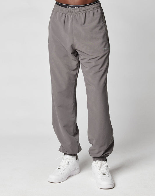 A person wears LIVE FAST DIE YOUNG LFDY Trackpants with Elastikbund and white sneakers, standing against a plain white background. Only the lower half is visible, highlighting the pants casual Regular Fit.