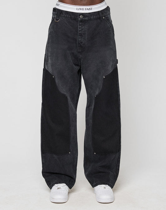 A person showcases the Carpenter Look in loose-fitting black LFDY Carpenter Pants by LIVE FAST DIE YOUNG, with large dark thigh patches, white sneakers, and a visible LIVE EAST waistband against a plain light background.
