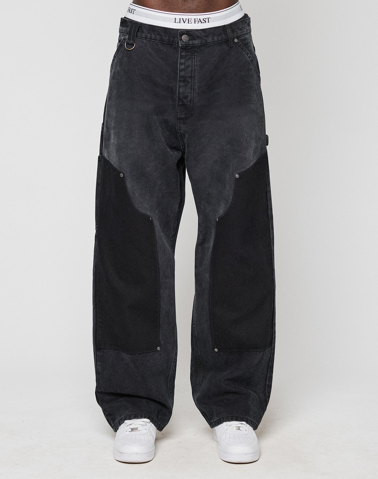 A person showcases the Carpenter Look in loose-fitting black LFDY Carpenter Pants by LIVE FAST DIE YOUNG, with large dark thigh patches, white sneakers, and a visible LIVE EAST waistband against a plain light background.