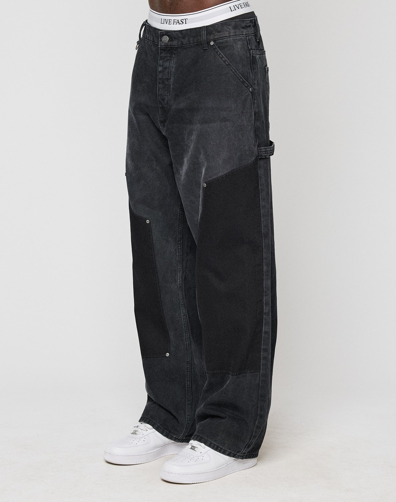 A person models the classic Carpenter Look in baggy-fit, black LFDY Carpenter Pants by LIVE FAST DIE YOUNG, featuring large dark knee patches and a LIVE FAST waistband, paired with white sneakers. The background is plain and light-colored. | model