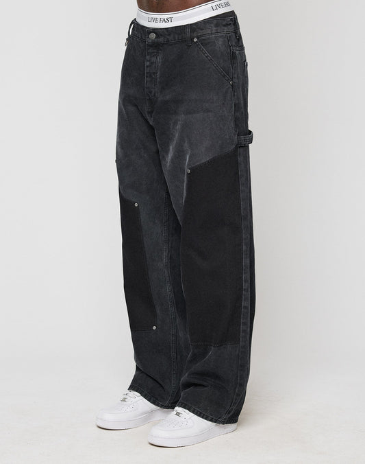 A person models the classic Carpenter Look in baggy-fit, black LFDY Carpenter Pants by LIVE FAST DIE YOUNG, featuring large dark knee patches and a LIVE FAST waistband, paired with white sneakers. The background is plain and light-colored. | model