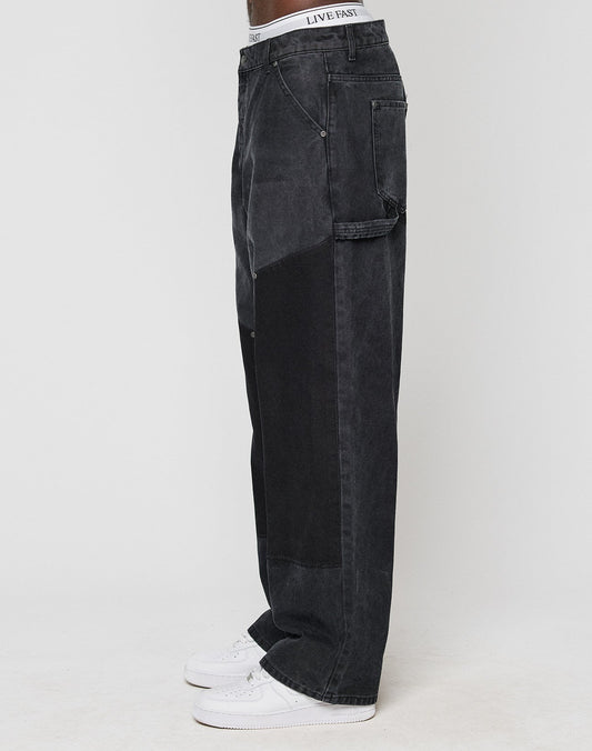 Model wears LFDY Carpenter Pants by LIVE FAST DIE YOUNG—baggy fit black denim with carpenter style, side pocket, and white waistband showing. Paired with white sneakers, standing sideways against a plain background. Made from 100% Baumwolle.
