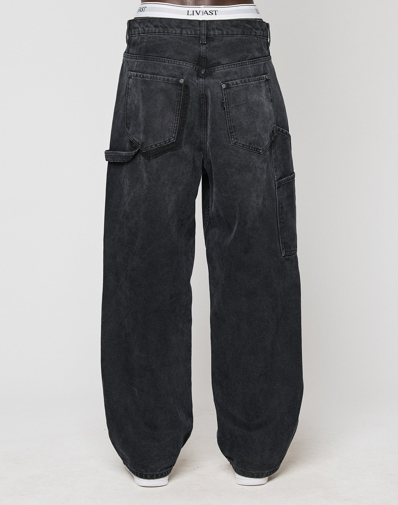 A person wearing LFDY Carpenter Pants by LIVE FAST DIE YOUNG, featuring a loose baggy fit in faded black with large back pockets and a tool pocket, shown from the back against a plain white background.