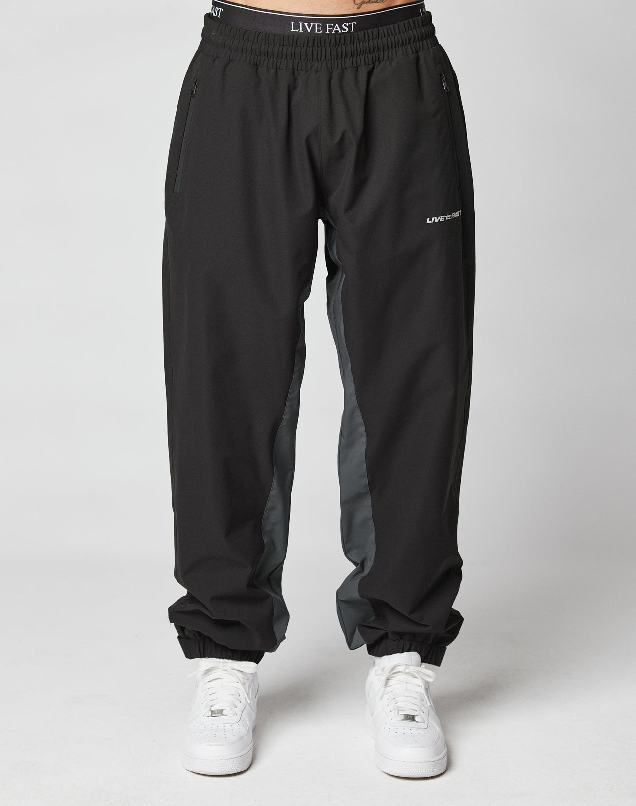 A person wears LIVE FAST DIE YOUNG Basic Shell Pants—black joggers with gray side panels, elastic drawstring waist reading “LIVE FAST,” zippered side pockets, and a reflective logo on the left thigh. Styled with white sneakers; plain white background.