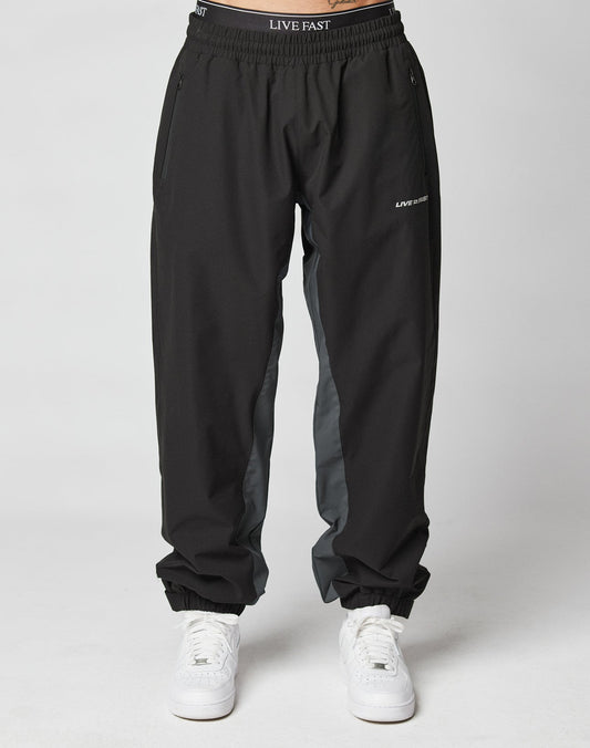 A person wears LIVE FAST DIE YOUNG Basic Shell Pants—black joggers with gray side panels, elastic drawstring waist reading “LIVE FAST,” zippered side pockets, and a reflective logo on the left thigh. Styled with white sneakers; plain white background.