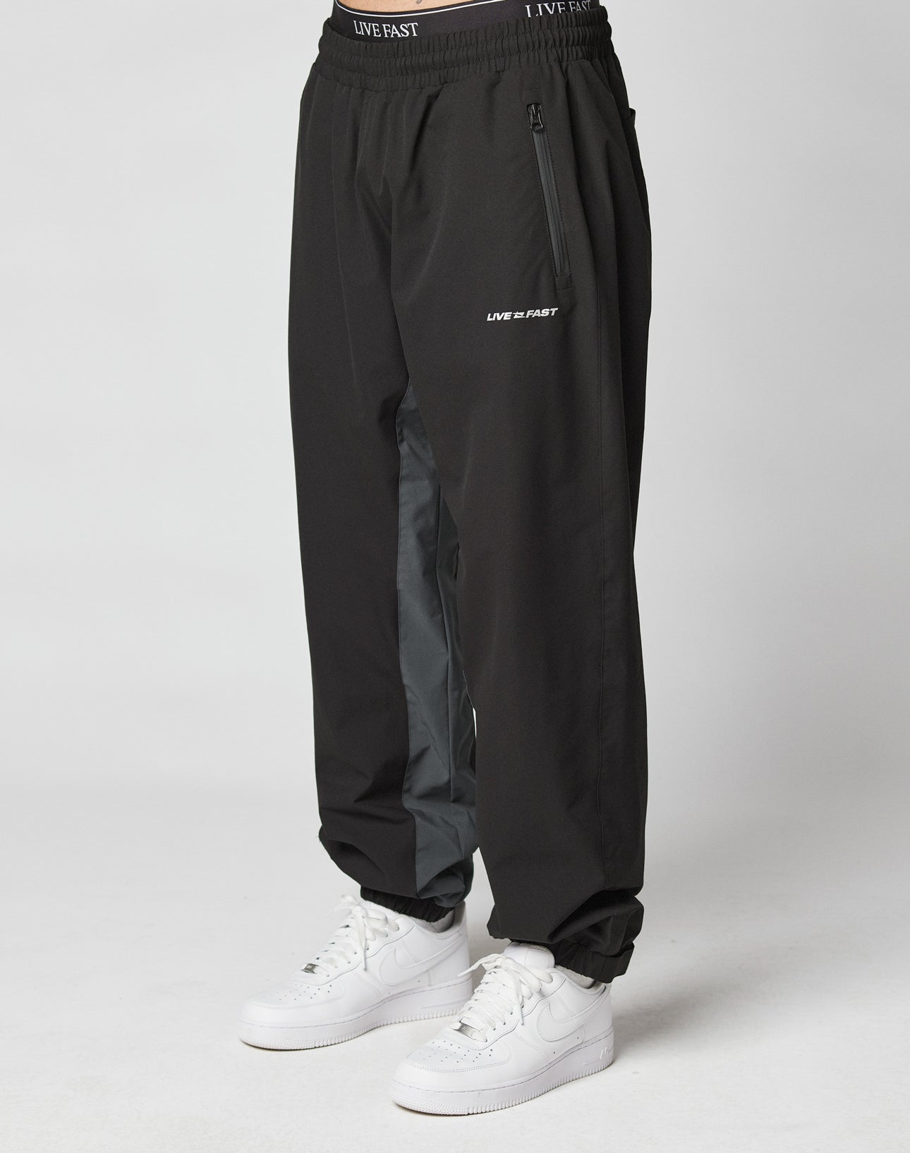A person wears LIVE FAST DIE YOUNG Basic Shell Pants with an elastic waistband and drawstring, “LIVE FAST” reflective print near the left pocket, a gray inner leg stripe, and white sneakers, standing against a plain light background. | model