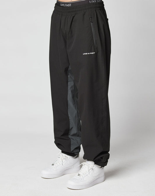 A person wears LIVE FAST DIE YOUNG Basic Shell Pants with an elastic waistband and drawstring, “LIVE FAST” reflective print near the left pocket, a gray inner leg stripe, and white sneakers, standing against a plain light background. | model