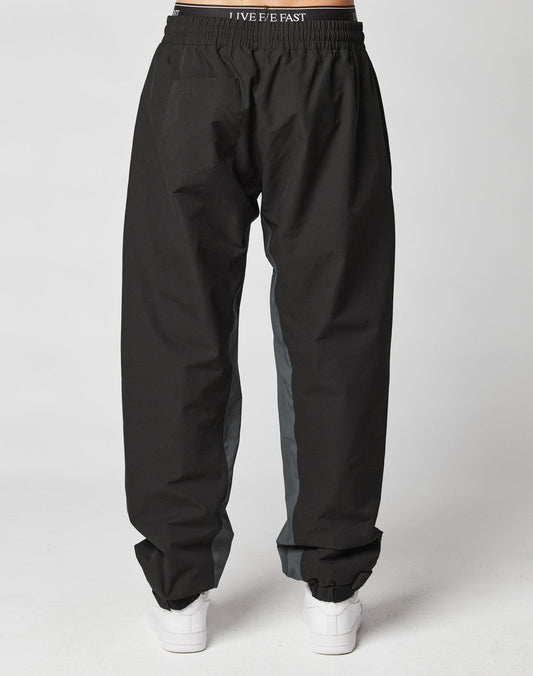 A person wearing LIVE FAST DIE YOUNG Basic Shell Pants, loose black with gray side panels and an elastic drawstring waistband, styled with white sneakers, photographed from the back against a plain white background.