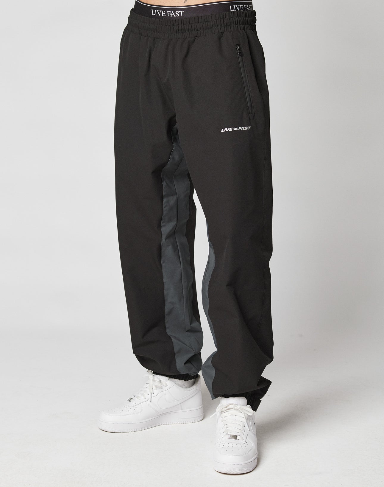 A person in black Basic Shell Pants by LIVE FAST DIE YOUNG, featuring gray side panels, a drawstring waist with LIVE FAST text, white sneakers, and a small LIVE FAST logo near the zipper pocket. The background is plain white.