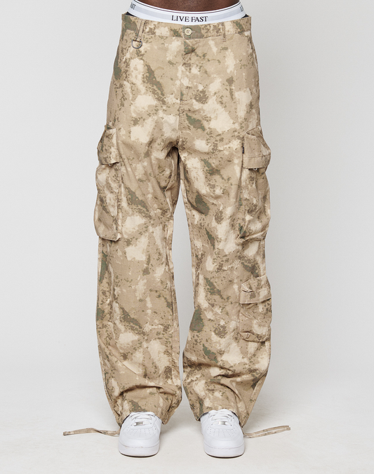 A person in LFDY Cargo pants by LIVE FAST DIE YOUNG—wide fit, tan camo print, 100% cotton, with cargo and side pockets—white LIVE FAST waistband visible, paired with white sneakers, against a plain white background.