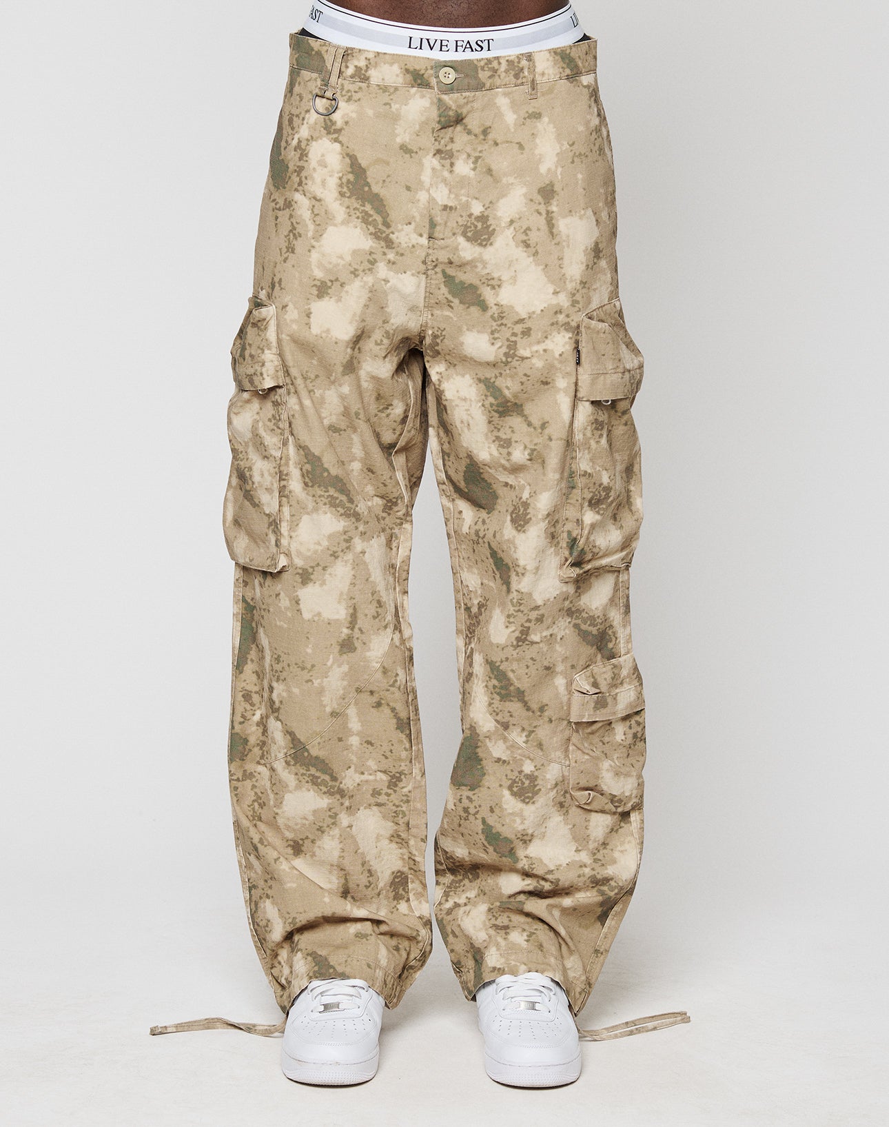 A person wears LFDY Cargo pants from LIVE FAST DIE YOUNG in wide fit tan camo with multiple cargo pockets and a visible LIVE FAST waistband. Made from 100% cotton, styled with white sneakers against a plain white background.