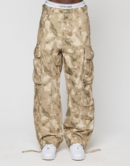 A person wears LFDY Cargo pants from LIVE FAST DIE YOUNG in wide fit tan camo with multiple cargo pockets and a visible LIVE FAST waistband. Made from 100% cotton, styled with white sneakers against a plain white background.