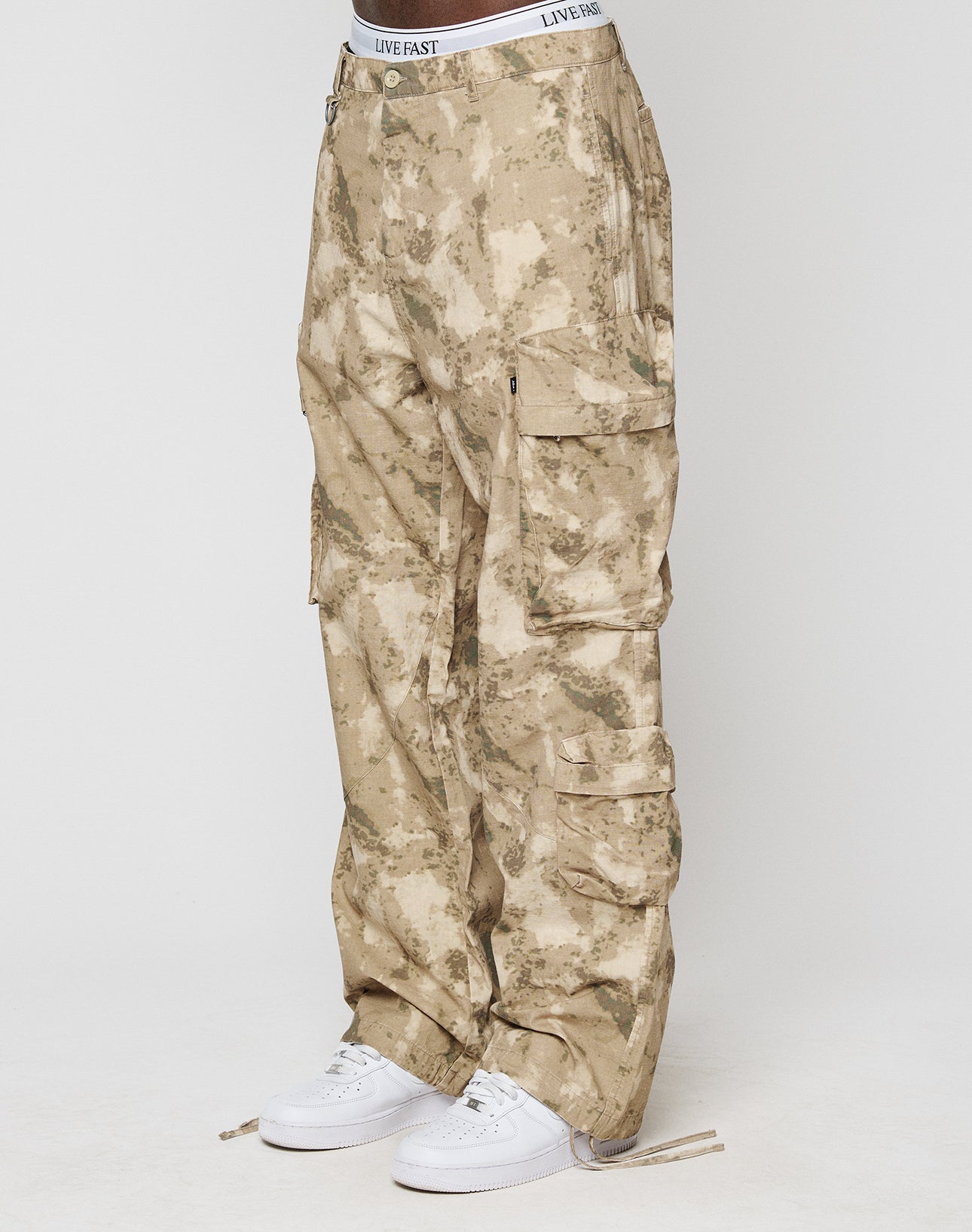 A person wears LFDY Cargo by LIVE FAST DIE YOUNG: loose-fitting tan camo cargo pants with Cargo Taschen, Wide Fit, and a white LIVE FAST waistband. Theyre paired with white sneakers. Made from 100% Baumwolle; backdrop is plain light gray.