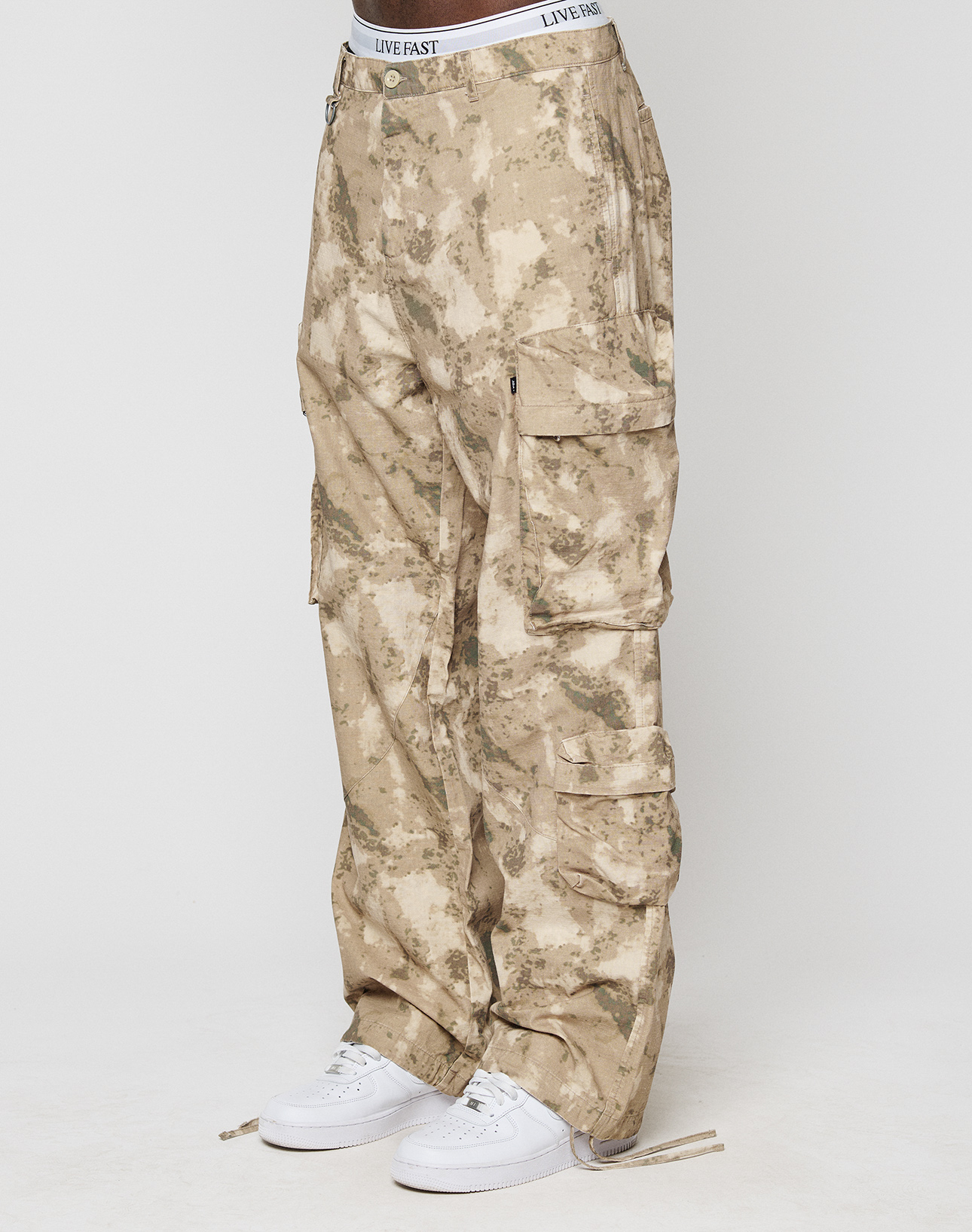 A person wears beige and tan camouflage LFDY Cargo pants from LIVE FAST DIE YOUNG, paired with white sneakers and a white LIVE FAST waistband, standing against a plain white background. | model