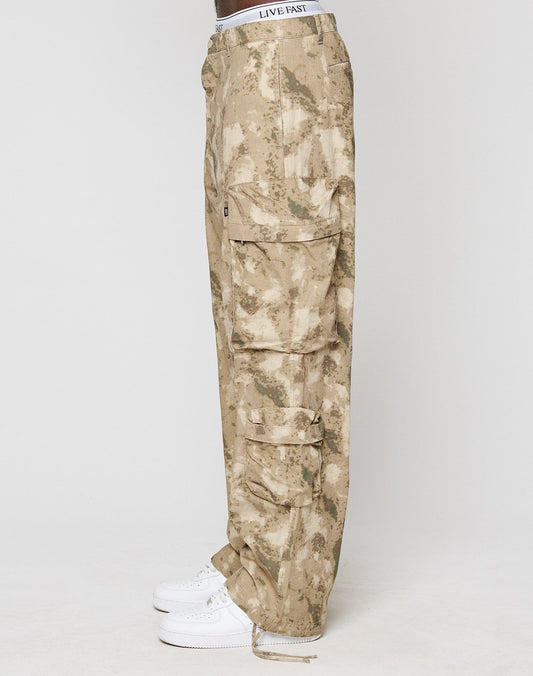 Side view of a person wearing LFDY Cargo Wide Fit pants from LIVE FAST DIE YOUNG in beige camouflage (100% Baumwolle), featuring Cargo Taschen, paired with white sneakers and a white LIVE FAST waistband, against a plain white background.