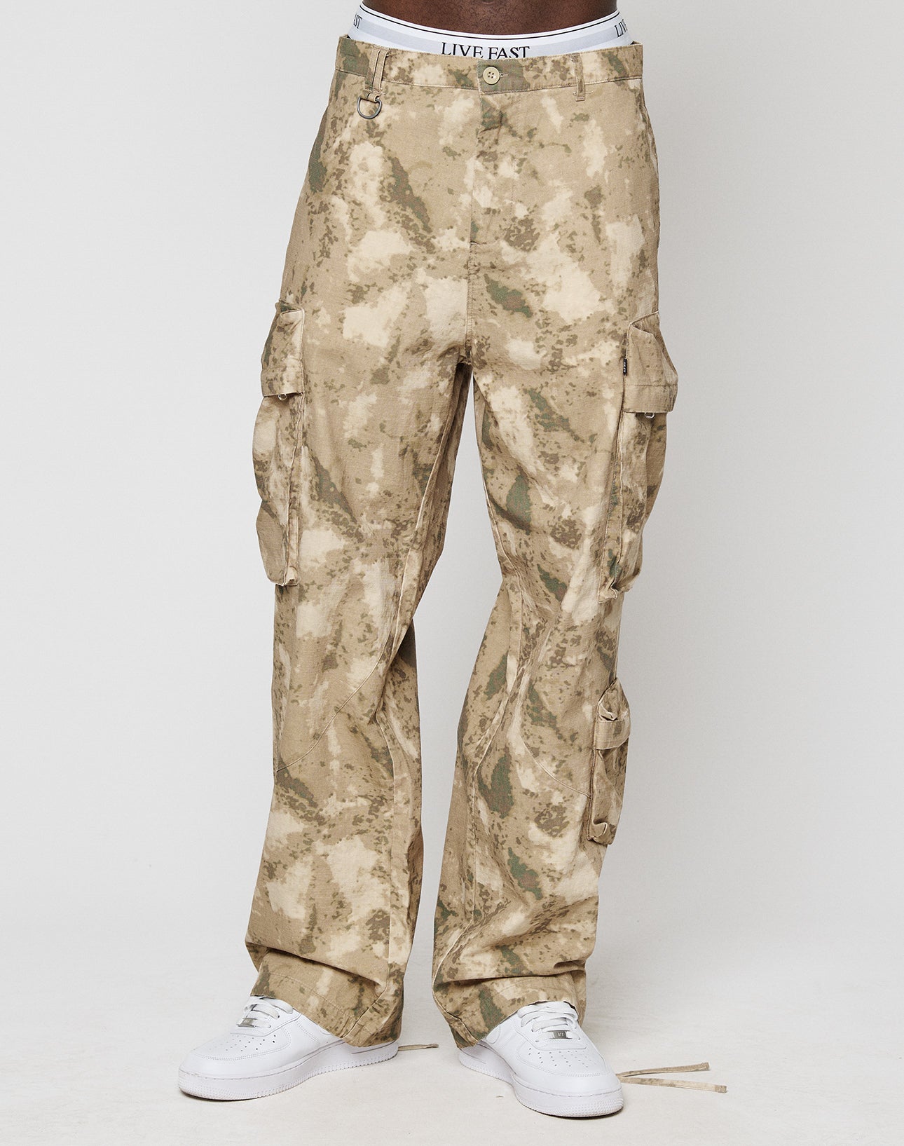 A person wearing beige LFDY Cargo pants by LIVE FAST DIE YOUNG with wide fit and cargo pockets, paired with white sneakers and a visible white waistband with “LIVE FAST” text, stands against a plain light background.