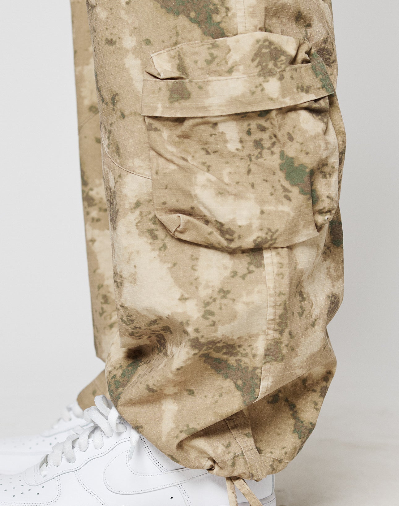 A close-up of a lower leg wearing LFDY Cargo pants by LIVE FAST DIE YOUNG in beige and green camo with großen Cargo Taschen, featuring a large side pocket, paired with white sneakers.