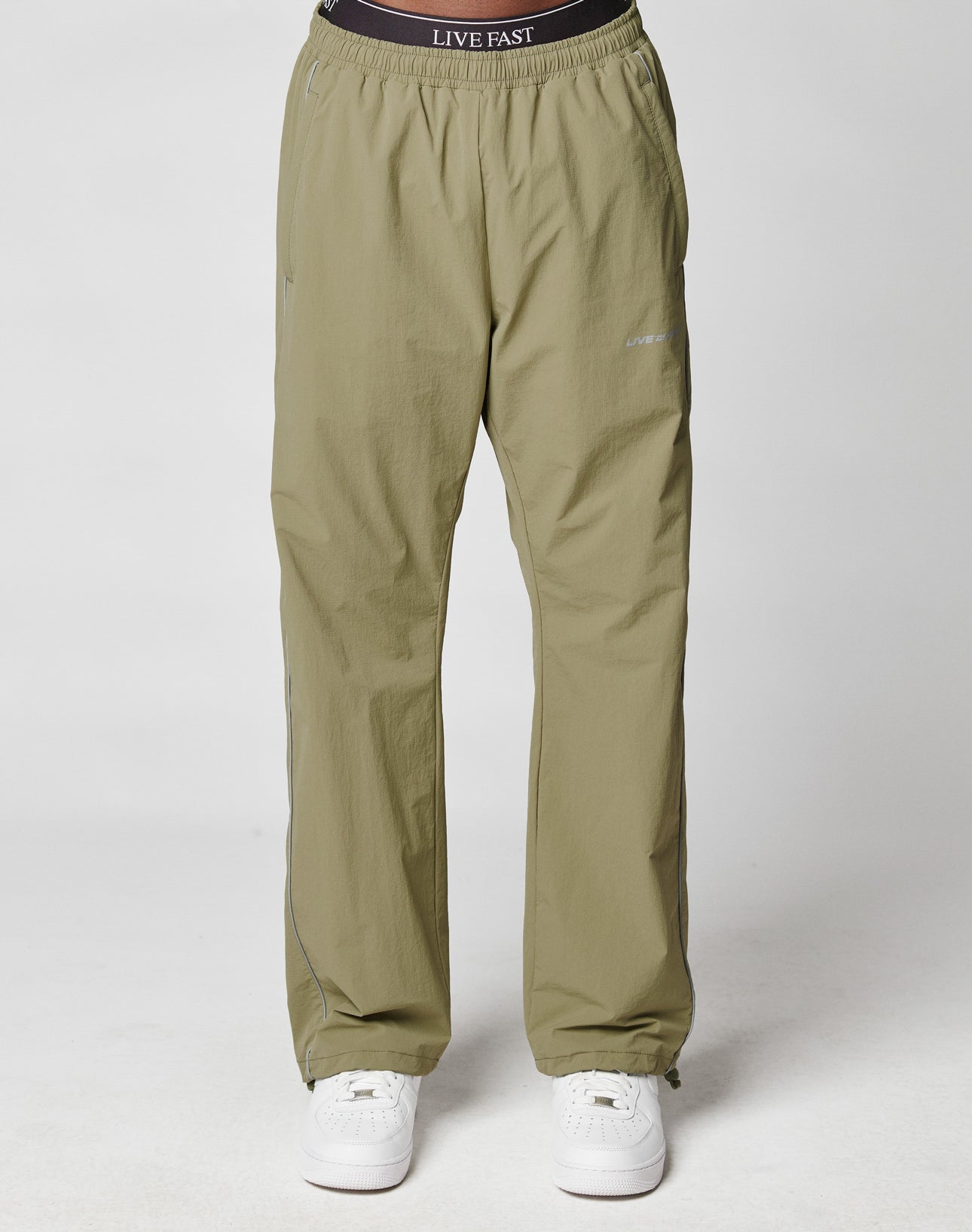 A person in light olive green, wide fit Live Fast Trackpants by LIVE FAST DIE YOUNG, featuring an Elastik-Kordelzug waistband and branding, is paired with white sneakers against a plain white background.
