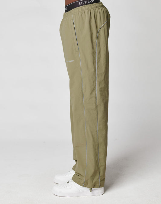 A person stands sideways against a plain white background wearing LIVE FAST DIE YOUNG’s olive green Live Fast Trackpants with white piping, elastic drawcord, side pockets, white sneakers, and a visible black LIVE FAST waistband.