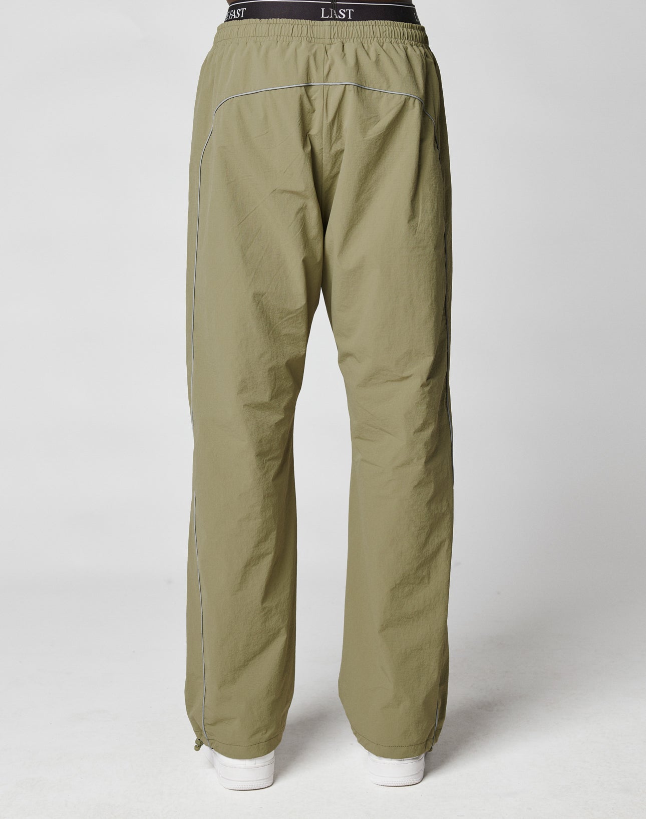 A person wearing LIVE FAST DIE YOUNG Live Fast Trackpants in light olive green wide fit with elastic drawcord waistband labeled LISI, reflective logo print, and white piping, stands back to camera against a plain background.