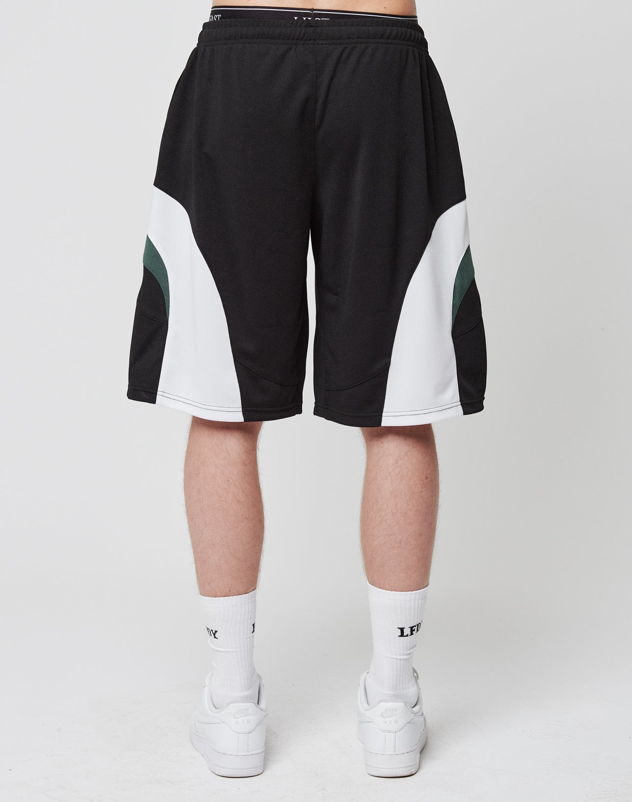 A person faces away, wearing LIVE FAST DIE YOUNG Basic Trackshorts—black, oversize fit with white and green accents and an elastic waistband—paired with white crew socks and sneakers against a plain white background.