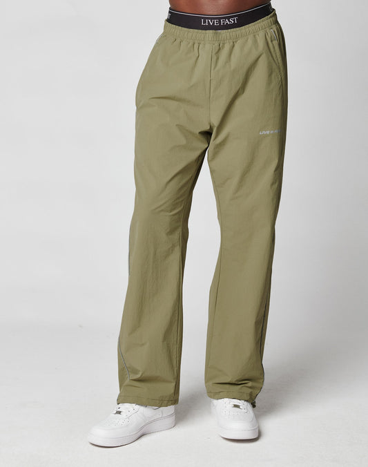 A person wears LIVE FAST DIE YOUNG Live Fast Trackpants in light olive green with an Elastik-Kordelzug and LIVE FAST waistband text, paired with white sneakers, standing against a plain light background.