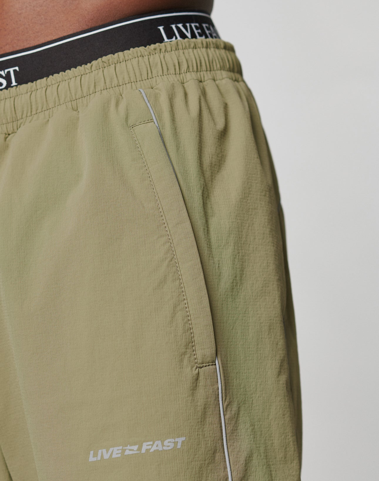 Close-up of a person wearing LIVE FAST DIE YOUNGs Live Fast Trackpants in light olive green. Wide Fit, white LIVE FAST near the pocket, black waistband with matching phrase, and an elastic drawcord for adjustable comfort.