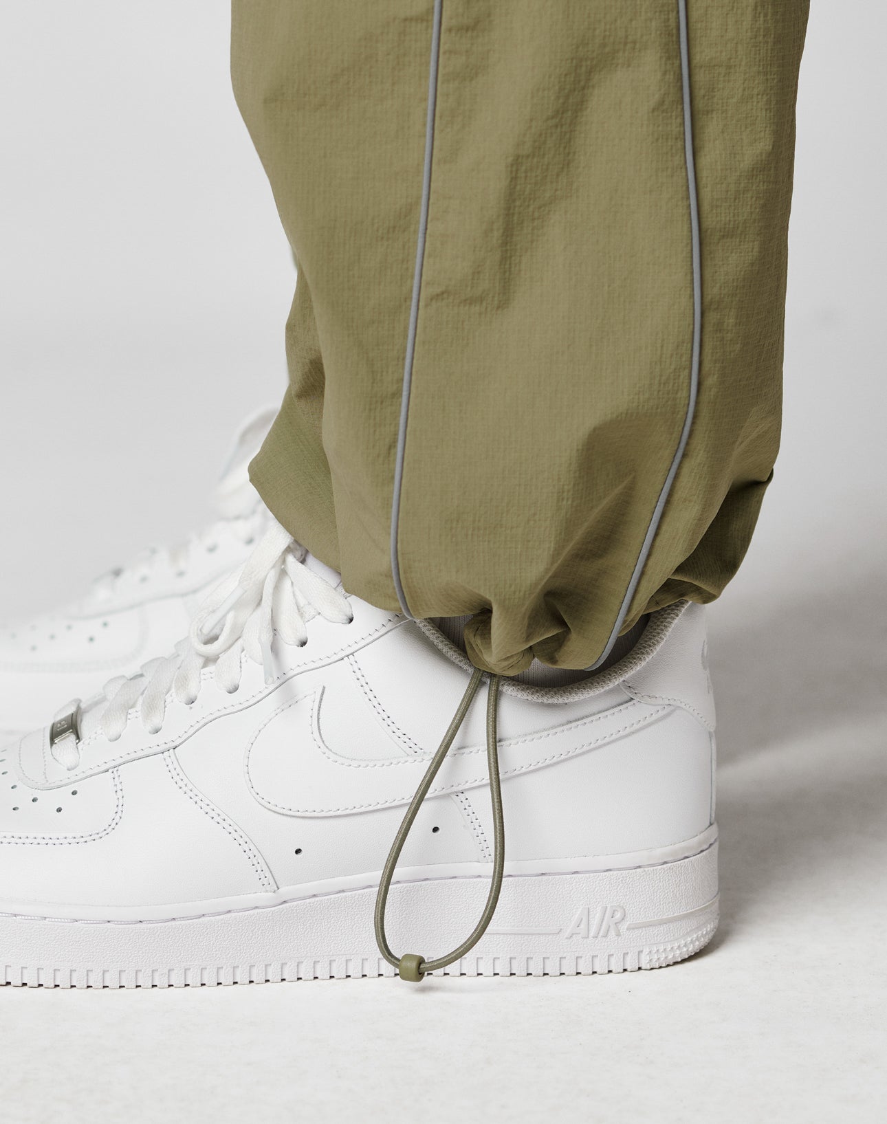 A close-up of a person wearing white Nike Air Force 1 sneakers and LIVE FAST DIE YOUNG Live Fast Trackpants in olive green with an elastic drawcord at the ankle and a reflective logo print, standing on a white surface.