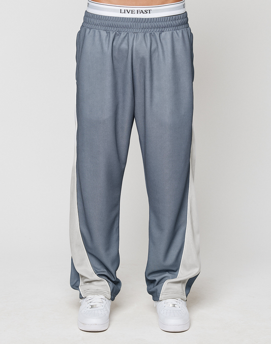 A person wears LIVE FAST DIE YOUNG's LFDY DY Trackpants in blue-gray with white side stripes and elastic waistband, paired with white sneakers and visible LIVE FAST underwear. Photo shows waist to feet against a plain background.