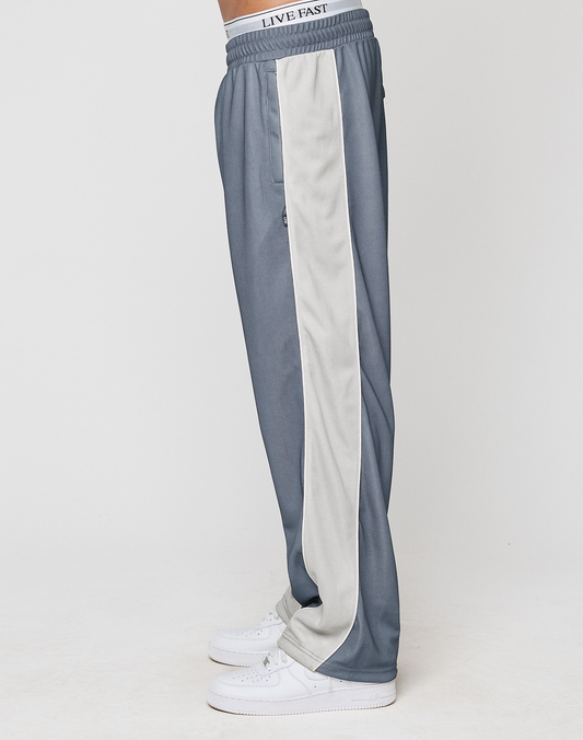 A person stands sideways wearing LFDY DY Trackpants by LIVE FAST DIE YOUNG, featuring a wide gray fit, light gray side stripe, elastic waistband with LIVE FAST label, and white sneakers against a plain white background.