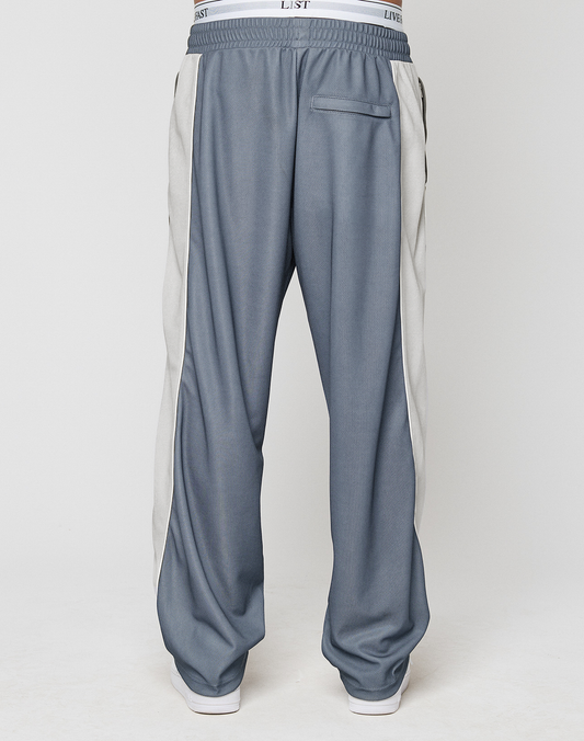 A person is shown from the back wearing LIVE FAST DIE YOUNG LFDY DY Trackpants in a wide fit blue design with light gray side panels and a visible white elastic waistband, standing against a plain light background.