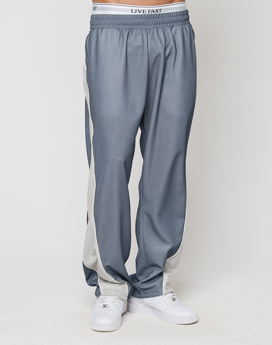 A person in LFDY DY Trackpants by LIVE FAST DIE YOUNG, featuring a blue-gray wide fit, white side stripes, and an elastic waistband with 'LIVE FAST' text, paired with white sneakers against a light background.