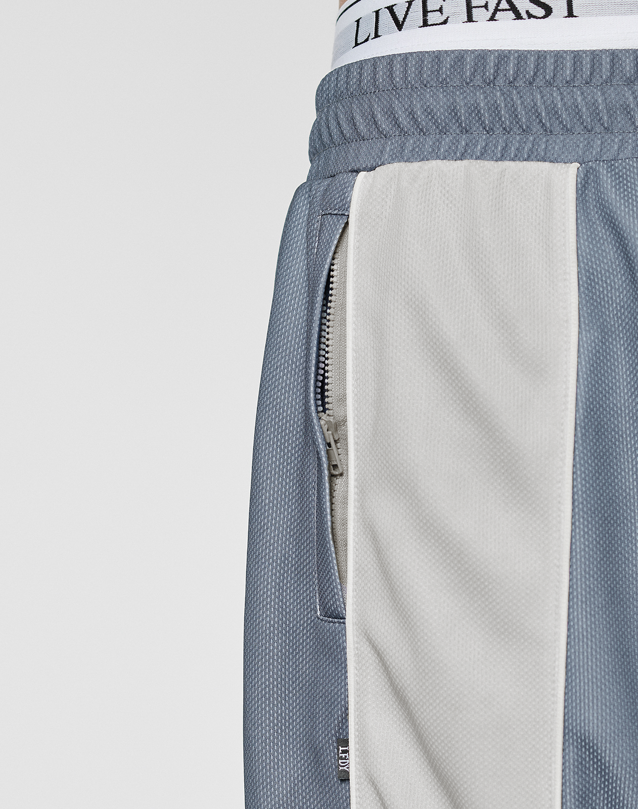 Close-up of a person wearing LFDY DY Trackpants by LIVE FAST DIE YOUNG, featuring blue and beige polyester fabric, a visible zipper pocket, and a white Elastikbund waistband with "LIVE FAST" printed on it.