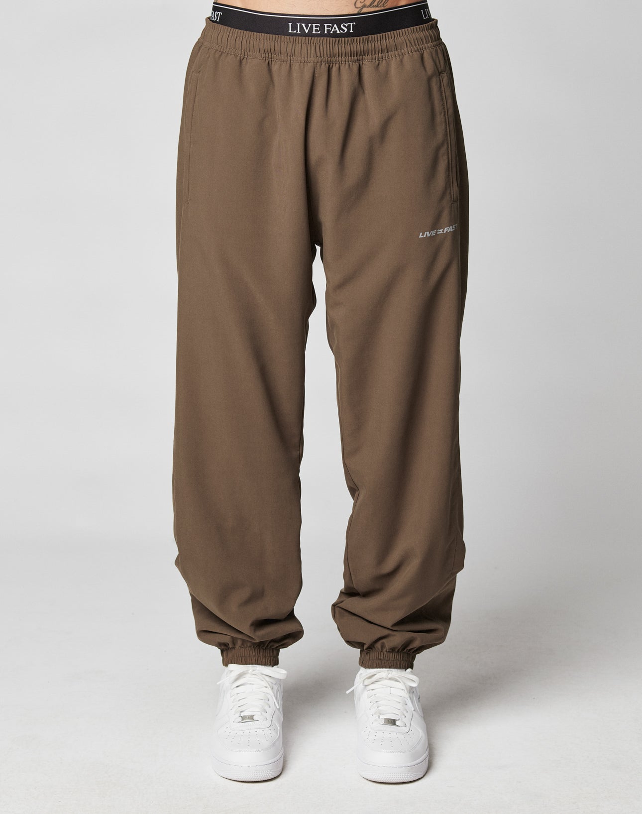 Someone stands against a plain light gray background wearing LFDY Trackpants by LIVE FAST DIE YOUNG, featuring an elastic waistband and reflective LIVE FAST print on the waistband and left thigh, paired with white sneakers.