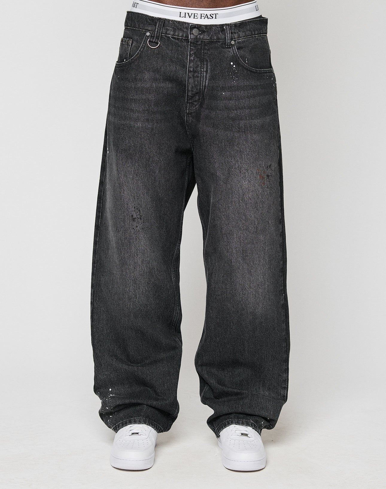 A person in LFDY Baggy Denim by LIVE FAST DIE YOUNG: faded black, baggy fit jeans with slight distressing and a straight leg, paired with white sneakers and a visible LIVE FAST waistband; upper body not shown.