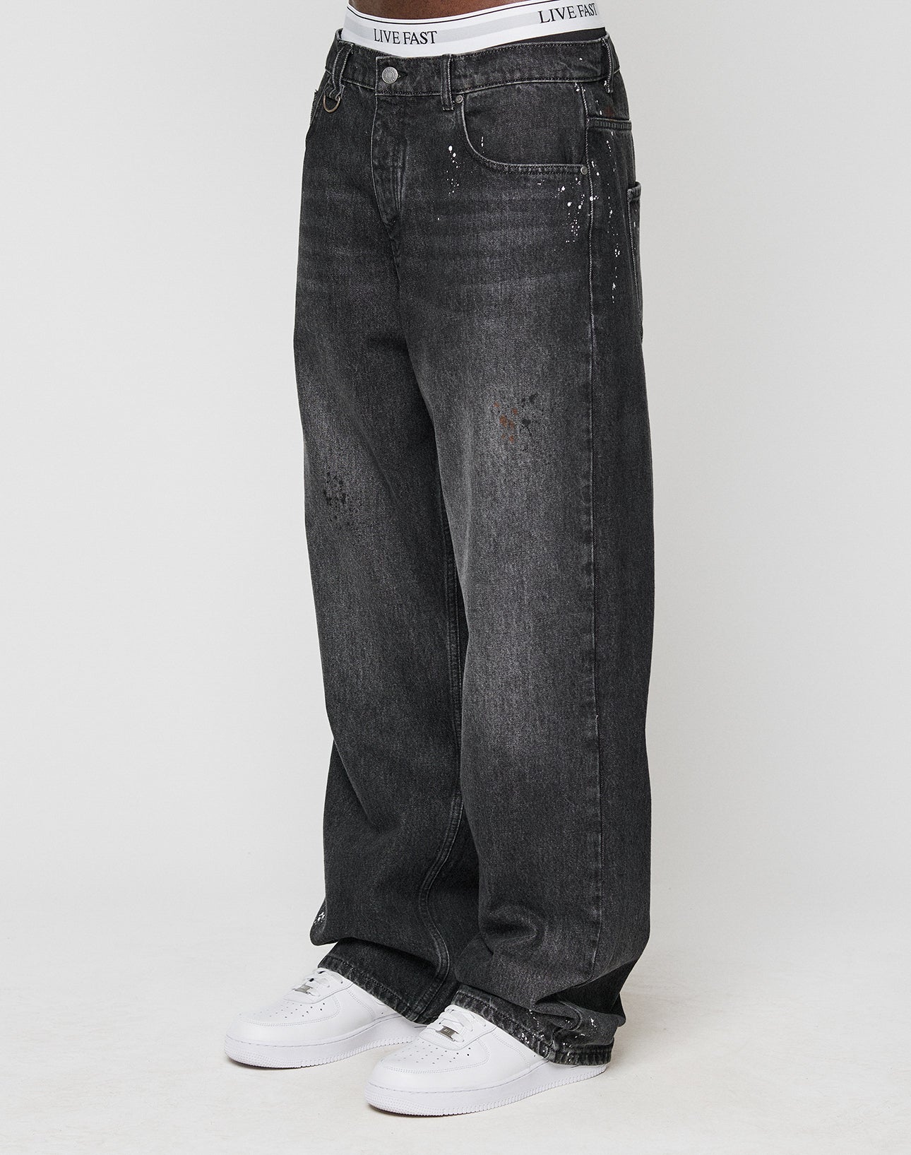 Someone wears the LFDY Baggy Denim by LIVE FAST DIE YOUNG—black jeans with wide straight legs, light fades, unique color splatters, subtle distressed details—paired with white sneakers and a visible LIVE FAST waistband.