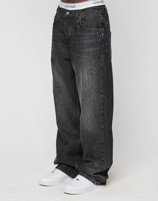 Someone wears the LFDY Baggy Denim by LIVE FAST DIE YOUNG—black jeans with wide straight legs, light fades, unique color splatters, subtle distressed details—paired with white sneakers and a visible LIVE FAST waistband. | model