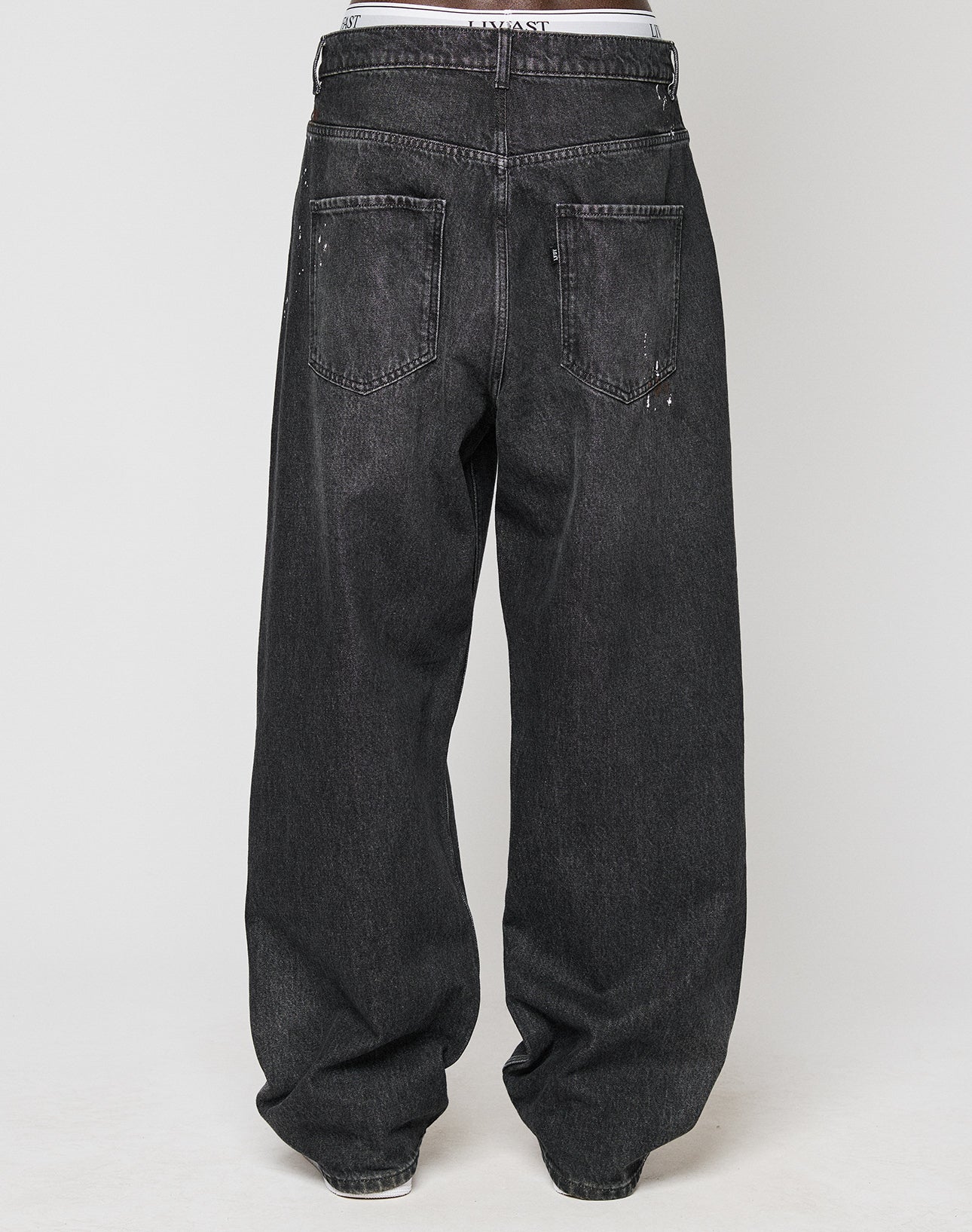 A person is seen from the back wearing LIVE FAST DIE YOUNGs LFDY Baggy Denim in dark gray, featuring a baggy fit, wide straight legs, visible back pockets, and a white waistband showing above the jeans.