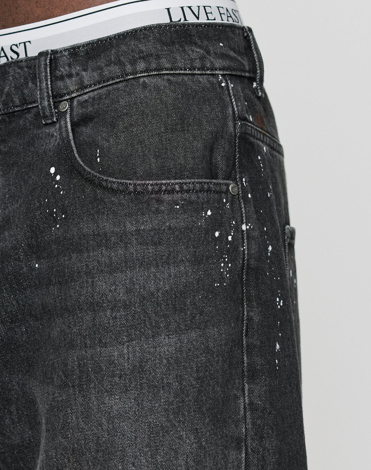 Close-up of a person wearing LFDY Baggy Denim by LIVE FAST DIE YOUNG, featuring individuelle Farbspritzer, with a visible white waistband that reads LIVE FAST in black letters.